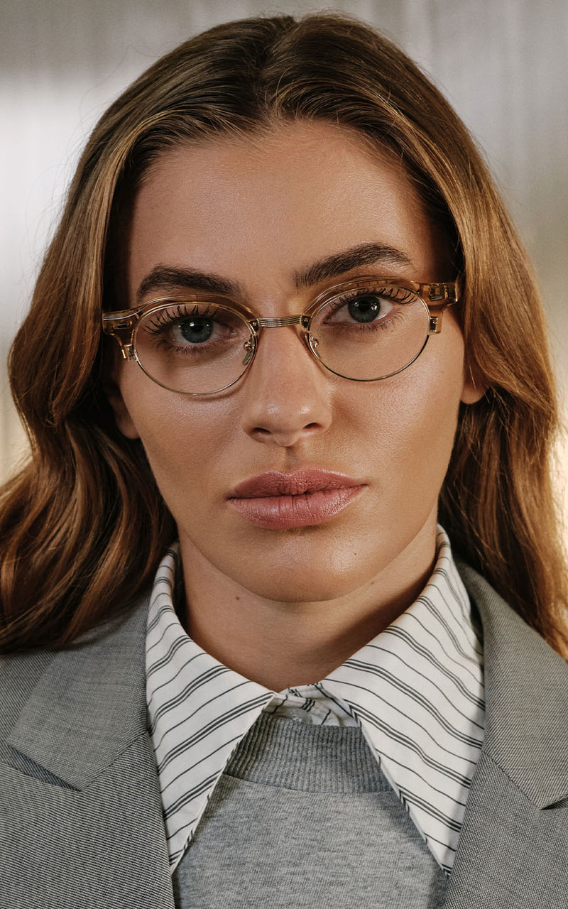 Mulher usando óculos de grau com armação clara e blazer cinza sobre camisa listrada 