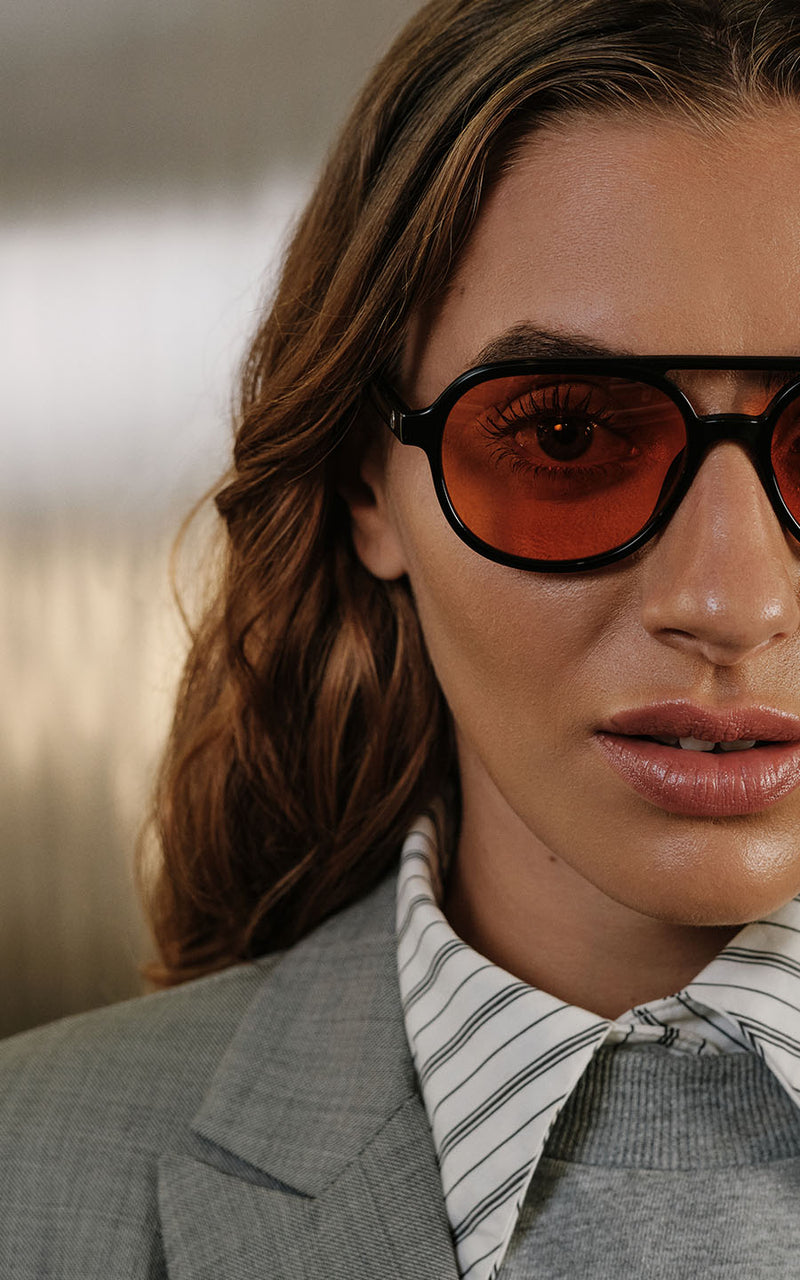 Mulher usa óculos de sol com lentes laranjas e armação preta, camisa social listrada e blazer cinza