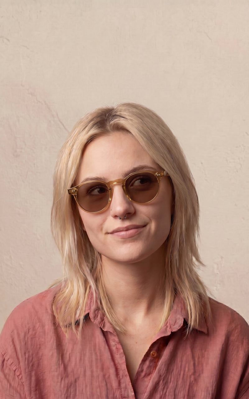 Mulher usando óculos de sol com armação redonda e clara, vestindo camisa rosa 