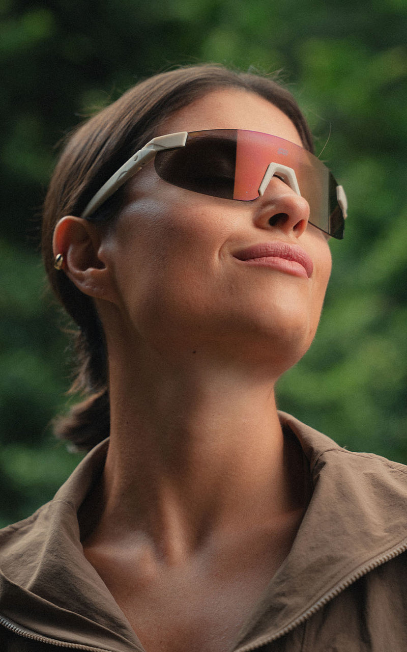 Mulher sorridente ao ar livre, usando óculos de sol esportivos com armação clara e lentes espelhadas