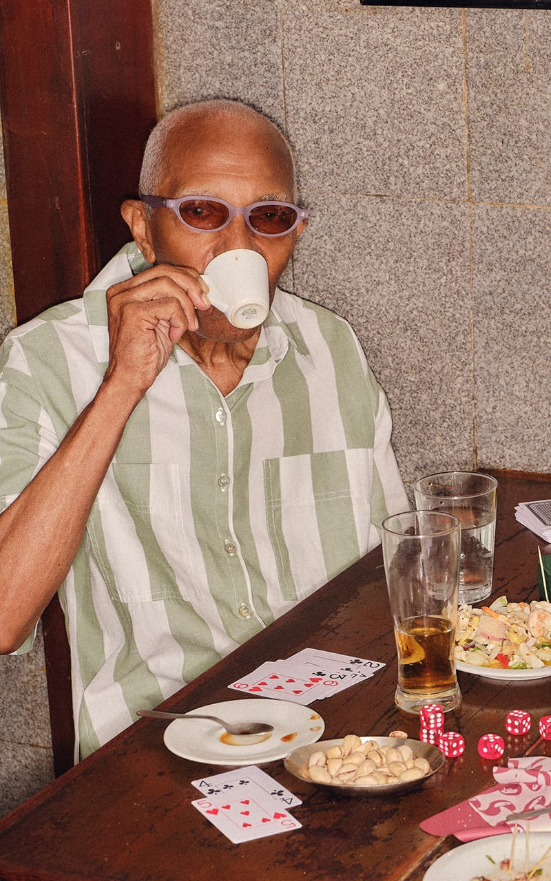 Homem de óculos e camisa listrada bebe café em uma mesa com cartas de baralho e petiscos 