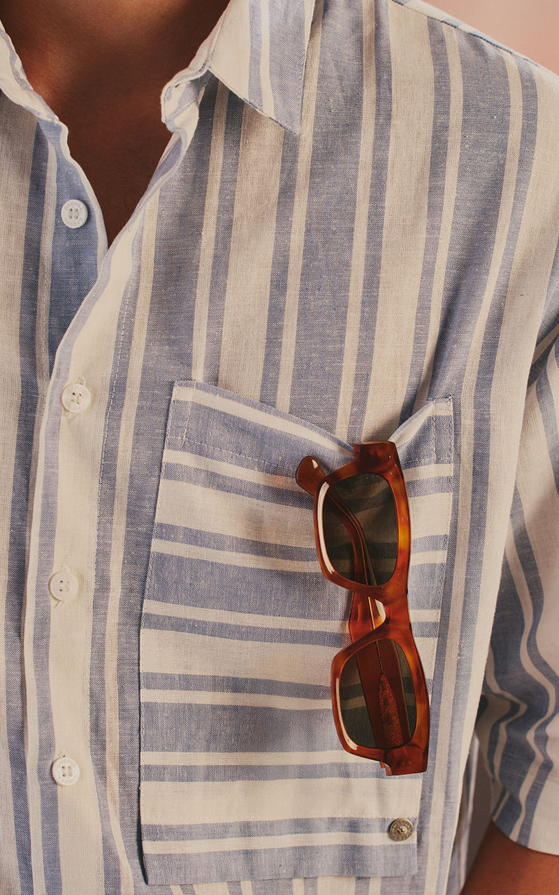 Homem vestindo camisa de linho listrada em azul e branco com óculos de sol no bolso frontal