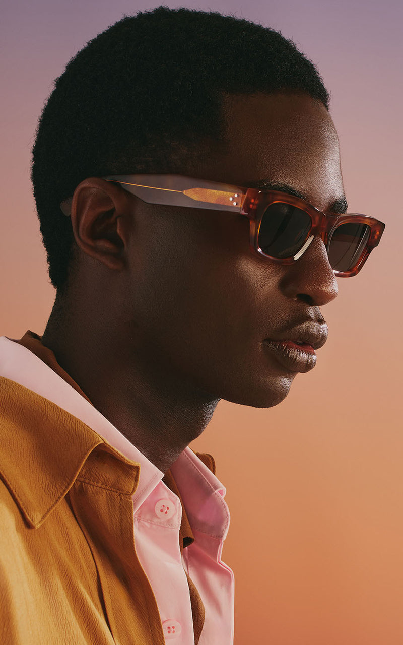 Homem usando óculos de sol com armação e camisa rosa contra fundo degradê