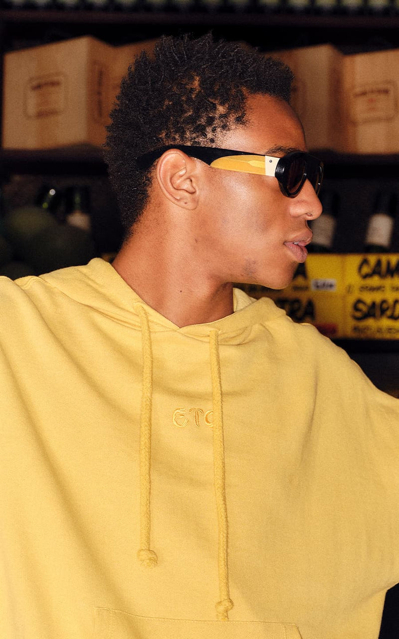 Jovem de perfil veste moletom amarelo com capuz e óculos de sol modernos em ambiente interno 