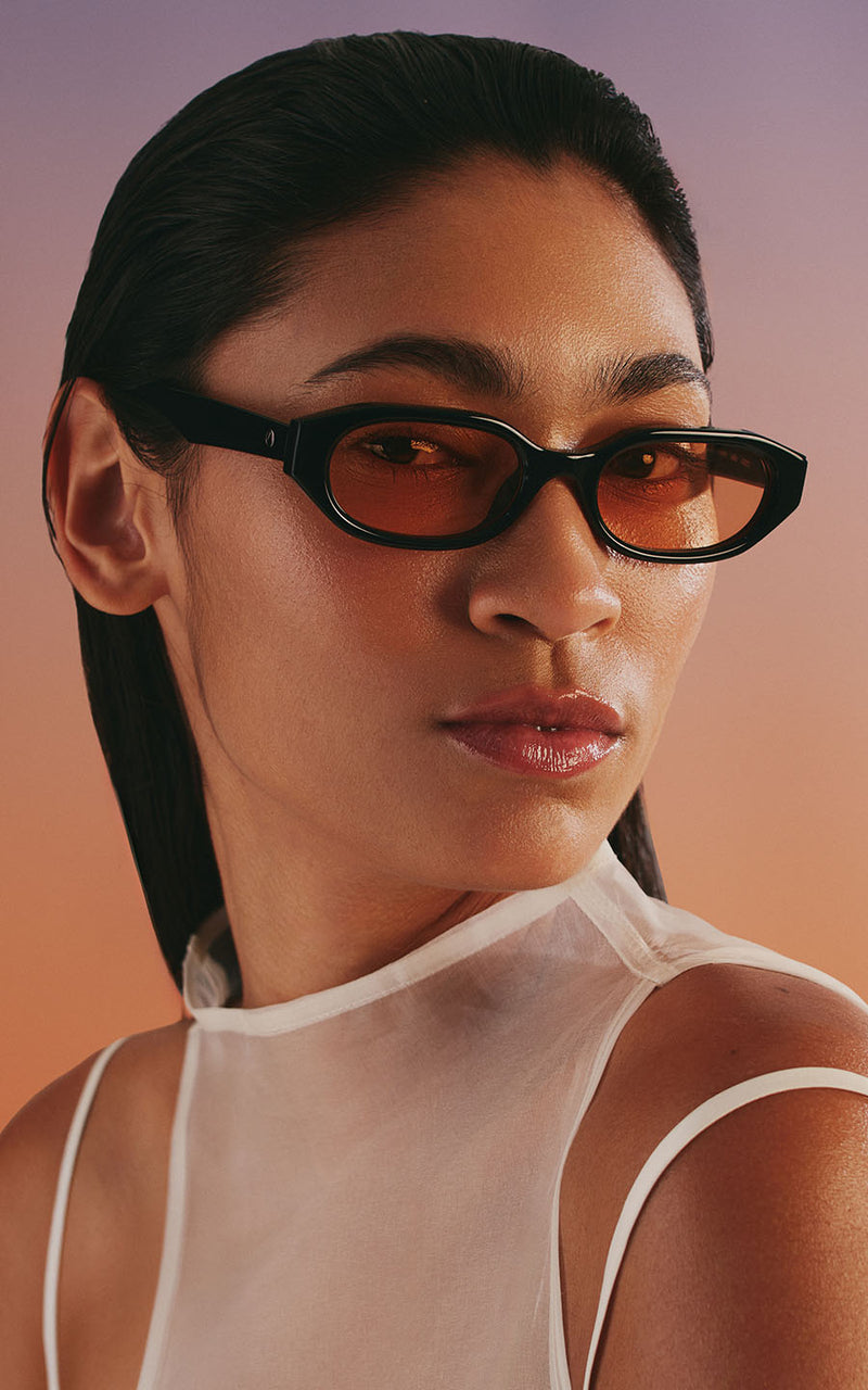 Mulher com blusa branca usa óculos de sol pretos com lentes alaranjadas contra fundo degradê