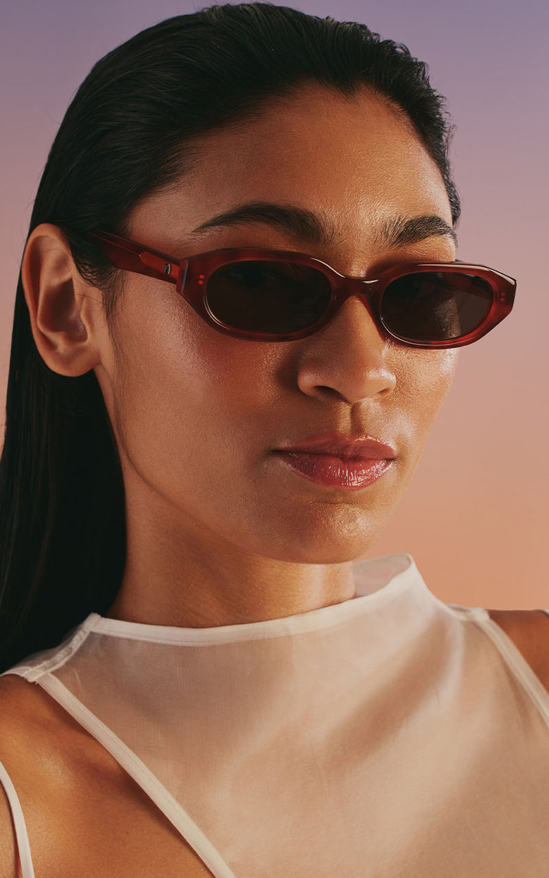 Mulher usando óculos de sol escuros e blusa branca transparente contra fundo degradê 